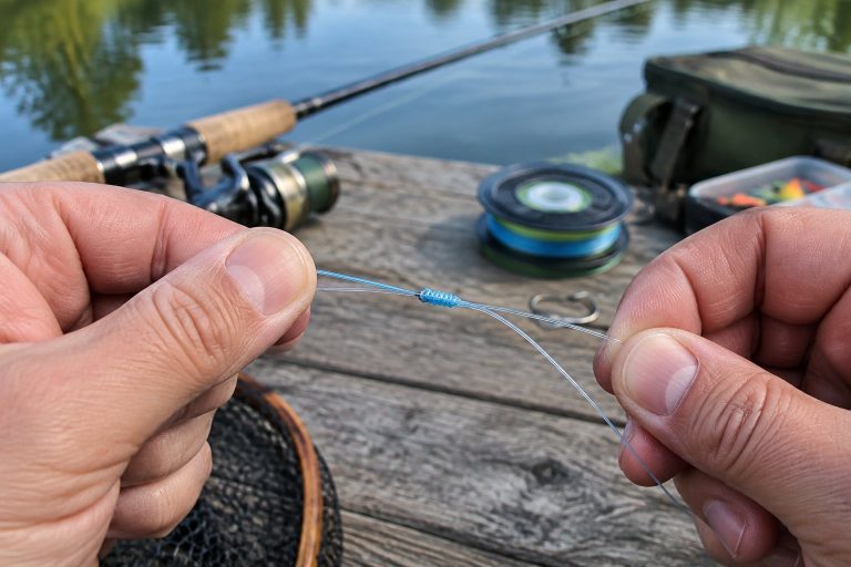 Close-up of an angler tying a blood knot in fishing line on a wooden dock beside calm water.